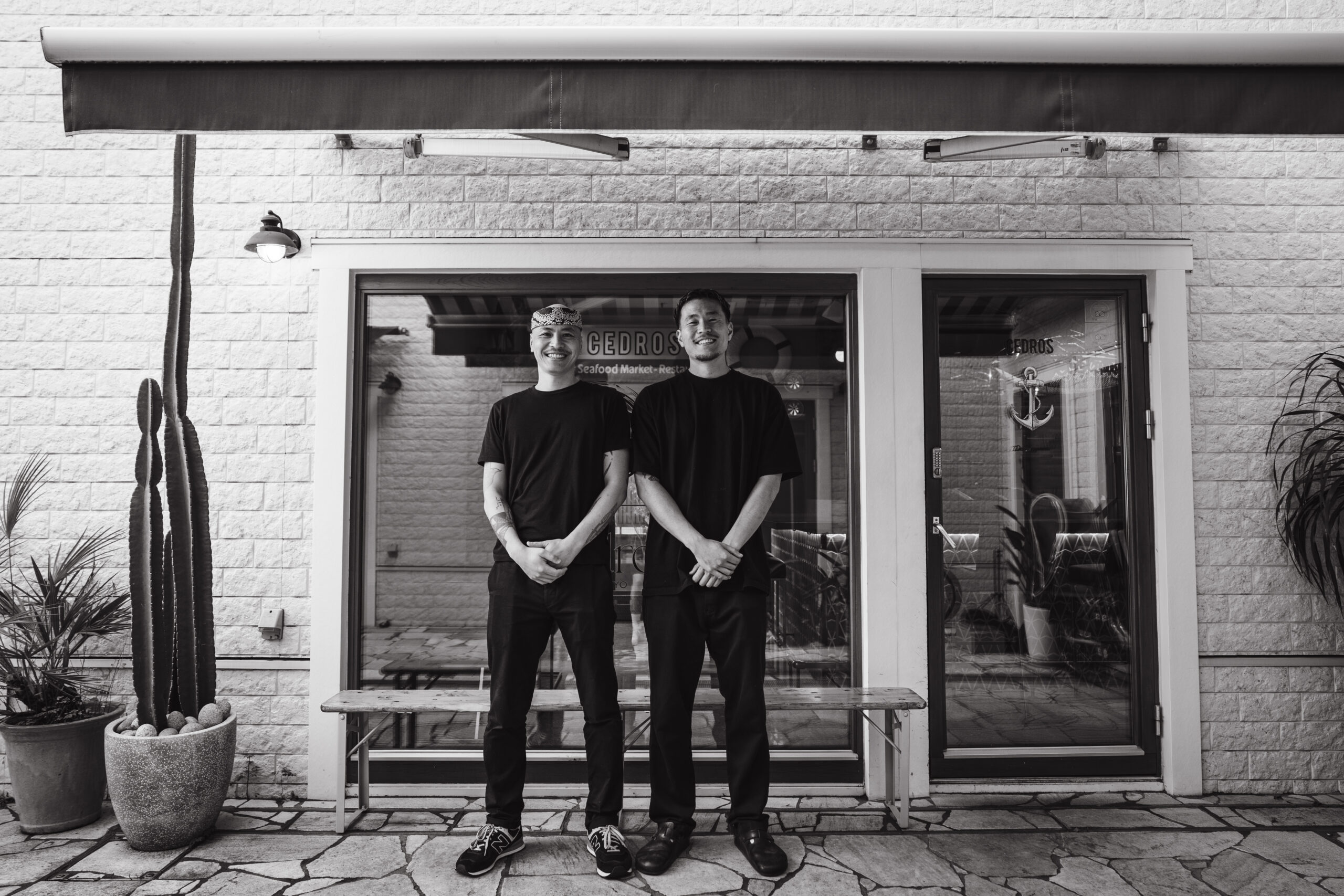 A black and white portrait of two male staff members from Cedros restaurant, standing side-by-side with hands clasped in front, outside the establishment with a large window displaying the restaurant's name. They are smiling confidently, conveying a sense of pride and hospitality.