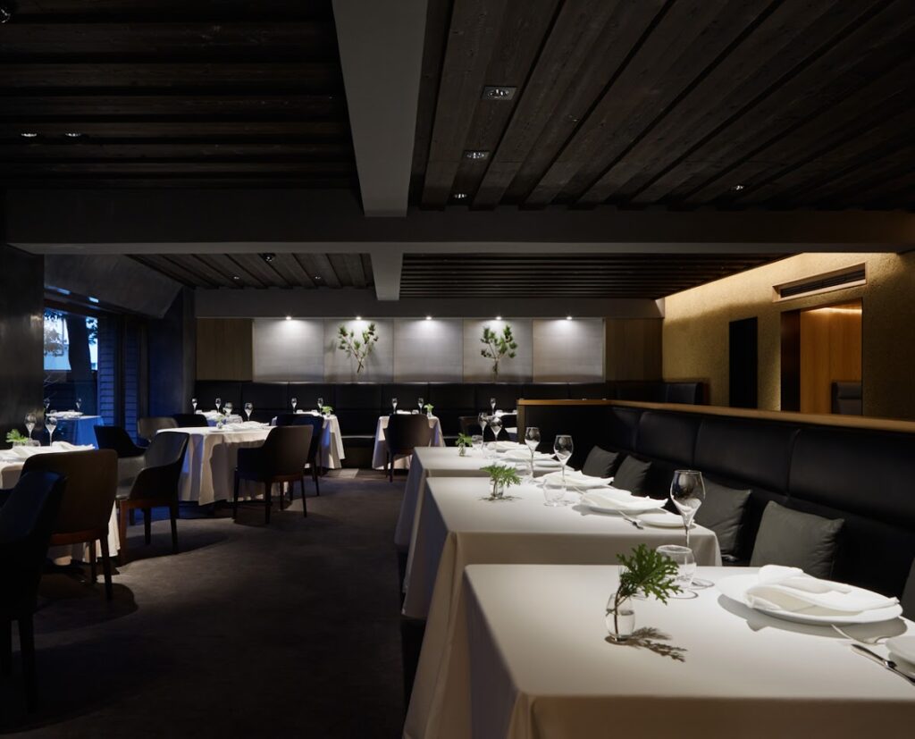 The sophisticated interior of a fine dining restaurant, L'Effervescence, featuring neatly set tables with white tablecloths and minimal green sprig decorations, plush seating, dark wood ceilings, and subtle lighting, creating an atmosphere of refined elegance.
