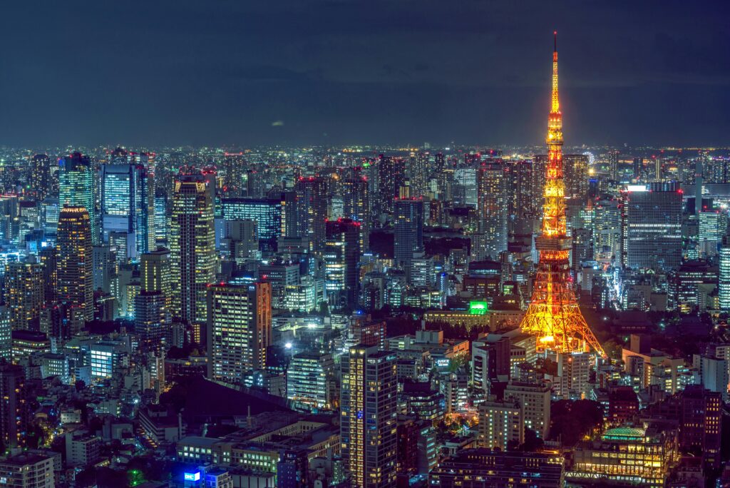 An expansive aerial view of the Tokyo cityscape at night, showcasing countless illuminated skyscrapers and buildings, with the prominently lit orange and white Tokyo Tower standing tall in the mid-ground, embodying the vibrant energy of Japan's capital after dark.
